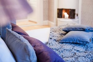Kakelugn i bakgrunden samt lila täcke och kuddar på sängen