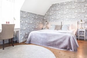 Rum39 Vallonhuset - lila överkast, blommiga tapeter, vit matta och beige skrivbordsstol. Trägolv.