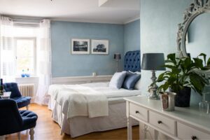 Hotellrum med ljusblåa väggar, blå sänggavel och vita gardiner, trägolv. En vit skänk med lampa och växt.