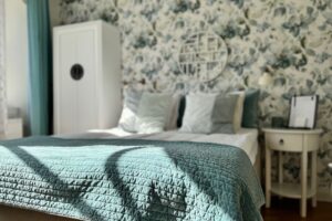 hotellrum - dubbelrum med turkost överkast och blå turkosa vita kuddan. Vitt sängbord och golvskåp. Vit tapet med blågröna bladmönster samt vitt väggdekoration med mönster.