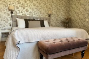 hotellrum med rosa överkast, en gammelrosa fotpall i plysch med knappar. Beige sänggavel och tapeter med vit bakgrund och bruna växtmönster.
