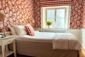 Hotellrum med en röd tapet med vita blommönster. Rutig veckgardin och vit blomkruka i fönstret med grön växt. Vita sängbord med lampor.