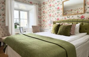 Dubbelrum med rosablommiga tapeter och säng med grönt överkast och kuddar (gröna och blommiga)