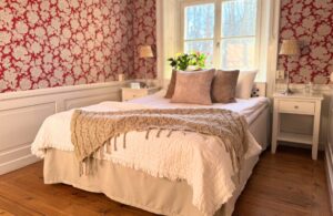 Hotellrum med röd tapet och vita blommor. grön växt i fönster, lila kuddar och stickat överkast.