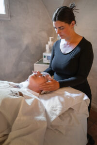 spaterapeut i svarta kläder och uppsatt hår gör ansiktsbehandling på liggand gäst i vit morgonrock.