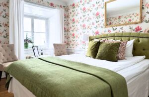 Dubbelrum med rosablommiga tapeter och säng med grönt överkast och kuddar (gröna och blommiga)