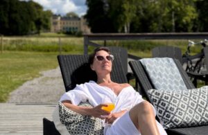 Kvinna i vit morgonrock ligger på en solbädd och håller ett glas juice. Slottet i bakgrunden